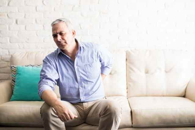 Reifer Mann mit grauem Haar, der Rückenschmerzen hat, während er zu Hause auf einer Couch sitzt