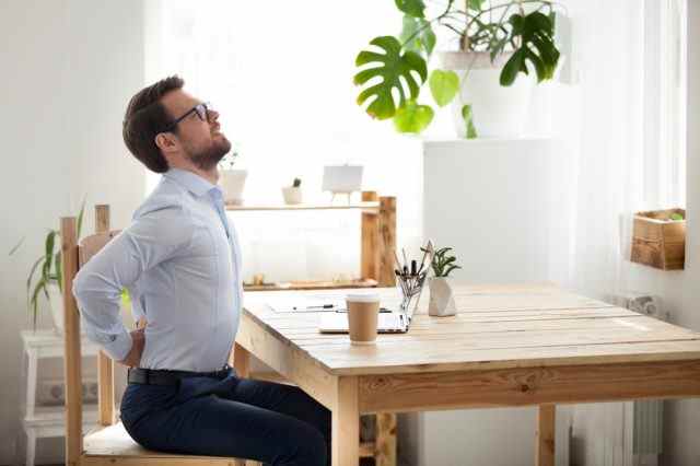Müde tausendjährige Büroangestellte, die sich auf dem Stuhl strecken, leiden unter langem Sitzen in falscher Haltung, männliche Angestellte haben Rückenschmerzen oder Wirbelsäulenkrämpfe, die in unbequemen Positionen arbeiten