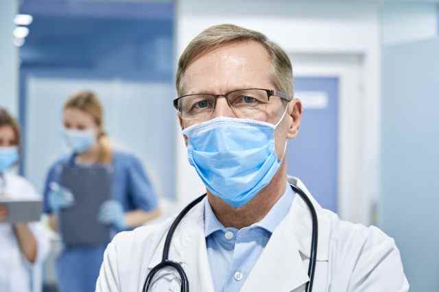 Reifer alter medizinischer Arzt mit weißem Mantel, Stethoskop, Brille und Gesichtsmaske, der in hospita.l steht und in die Kamera schaut