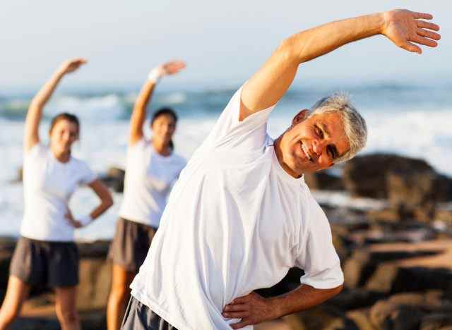 Mann mittleren Alters, der sich am Strand dehnt und Übungsgewohnheiten für Arthritis demonstriert