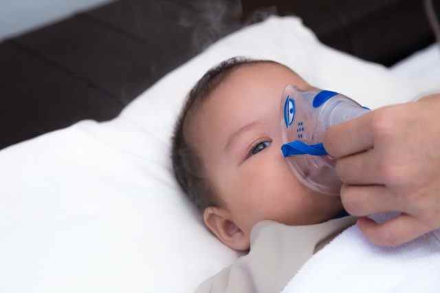 5 Monate altes Baby mit Respiratory-Syncytial-Virus, Inhalation von Medikamenten durch Inhalationsmaske, während es mit seinen müden Augen betrachtet
