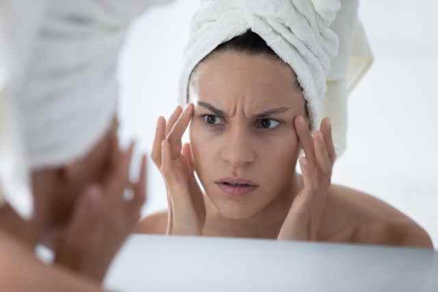 Unglückliche Frau mit weißem Badetuch, die die Haut nach dem Duschen überprüft, in den Spiegel schaut und die Gesichtshaut berührt.