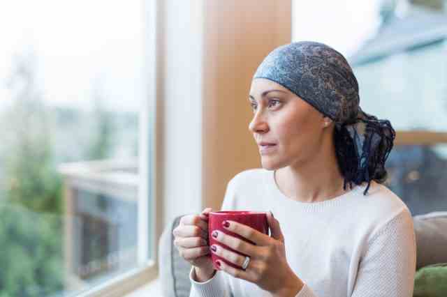 Frau in ihren 30ern sitzt mit einer Tasse Tee am Wohnzimmerfenster und schaut nachdenklich hinaus.  Sie ist eine Krebsüberlebende und trägt ein Kopftuch.