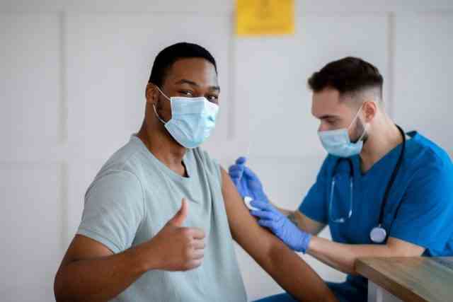 Afroamerikaner in antiviraler Maske, der während der Coronavirus-Impfung mit dem Daumen nach oben gestikuliert und die Covid-19-Immunisierung genehmigt