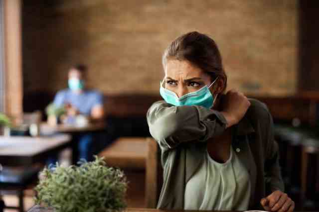 Frau mit Gesichtsmaske niest in den Ellbogen, während sie in einem Café sitzt.