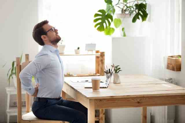 Müde tausendjährige Büroangestellte, die sich auf dem Stuhl strecken, leiden unter langem Sitzen in falscher Haltung, männliche Angestellte haben Rückenschmerzen oder Wirbelsäulenkrämpfe, die in unbequemen Positionen arbeiten