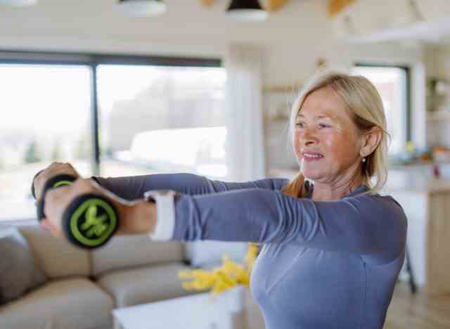 Eine fitte Seniorin zu Hause demonstriert Körperteile, die nach 60 mit Hanteln trainiert werden sollen