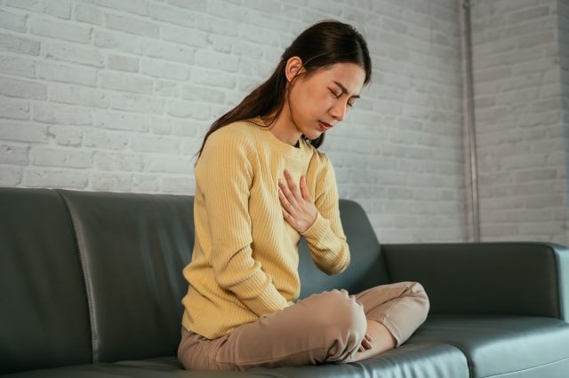Asiatische junge Frau fühlt sich unwohl, weil sie an Sodbrennen leidet, hält die Brust mit geschlossenen Augen und sitzt mit gefalteten Beinen auf der Couch zu Hause.
