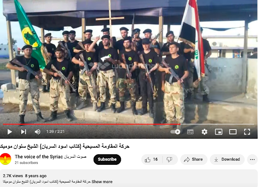 Dies ist ein Screenshot eines im Juli 2015 vom YouTube-Kanal „The Voice of the Syriac“ veröffentlichten Propagandavideos mit der gleichen Einstellung, das Salwan Momika zeigt, der eine Uniform trägt und ein Stück Papier in der Hand hält, scheinbar bereit, eine Spende zu geben Rede.
