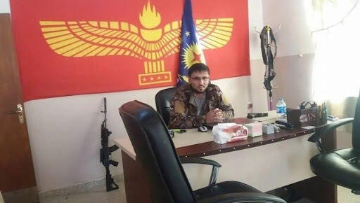 Dieses Foto von Salwan Momika, das vor seiner Ankunft in Schweden im April 2018 aufgenommen wurde, zeigt ihn in seinem Büro sitzend, eine Kalaschnikow in der Hand und vor der aramäischen (syrischen) Flagge sitzend, die eine ethnische Minderheitsgruppe darstellt.