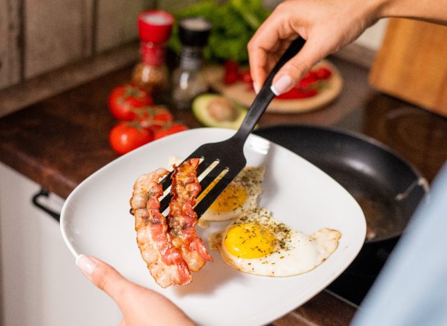 Nahaufnahme der Hände einer Frau, die Speck auf einen Teller mit Eiern neben dem Herd legt, Konzept von Lebensmitteln mit leeren Kalorien, die zu einer Gewichtszunahme führen