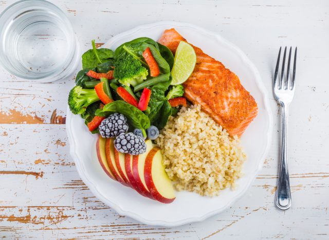 Portionierter Teller mit Fisch, Getreide, Gemüse, Obst und einem Wasserglas