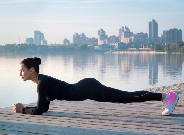 Frau macht Planken auf einem Pier am Wasser neben der Stadt, Konzept von Kraftübungen, um 10 Pfund in einem Monat abzunehmen