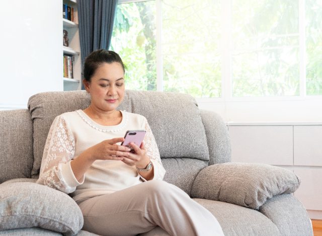Frau schreibt SMS auf ihrem Handy, während sie auf der Couch sitzt, anstatt Sport zu treiben, Trainingsgewohnheiten, die Ihren Körper zerstören