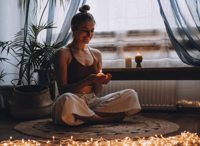 Glückliche Frau hält eine Kerze in der Hand und meditiert in ihrem Wohnzimmer, umgeben von Lichterketten