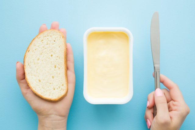 Frauenhände halten eine Scheibe Weißbrot und ein Messer.  Geöffnete Plastikpackung hellgelber Margarine auf pastellblauem Schreibtisch