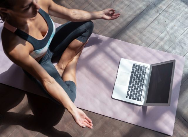 Frau in Yoga-Bekleidung meditiert zu Hause vor dem Laptop auf der Matte