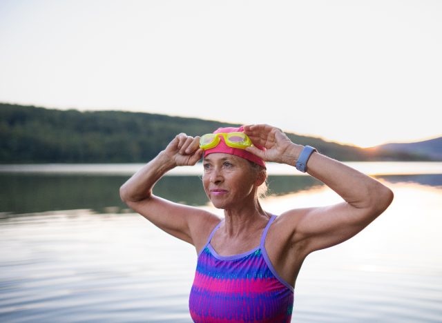 Reife, fitte Frau, die am See eine Schutzbrille aufsetzt und Cardio-Übungen vorführt, um Ihr Herz jung zu halten