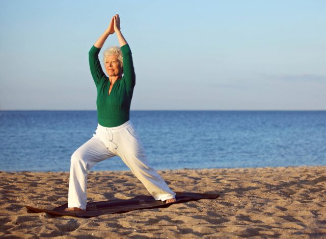 Reife Frau in grünem Langarm-Top und weißen Hosen macht Yoga zur goldenen Stunde am Strand