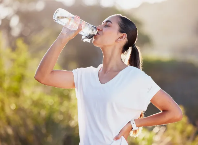 Glückliche brünette Frau in weißem T-Shirt, die an einem sonnigen Tag nach dem Training eine Wasserflasche im Freien trinkt